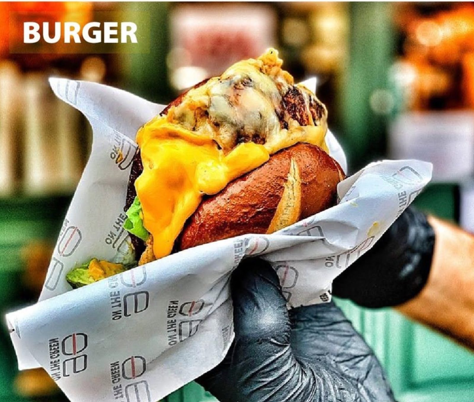In-the-wild moment — a cheeseburger wrapped in 90 On The Green branded greaseproof paper, gloved hand holding it, melted cheese spilling out the top