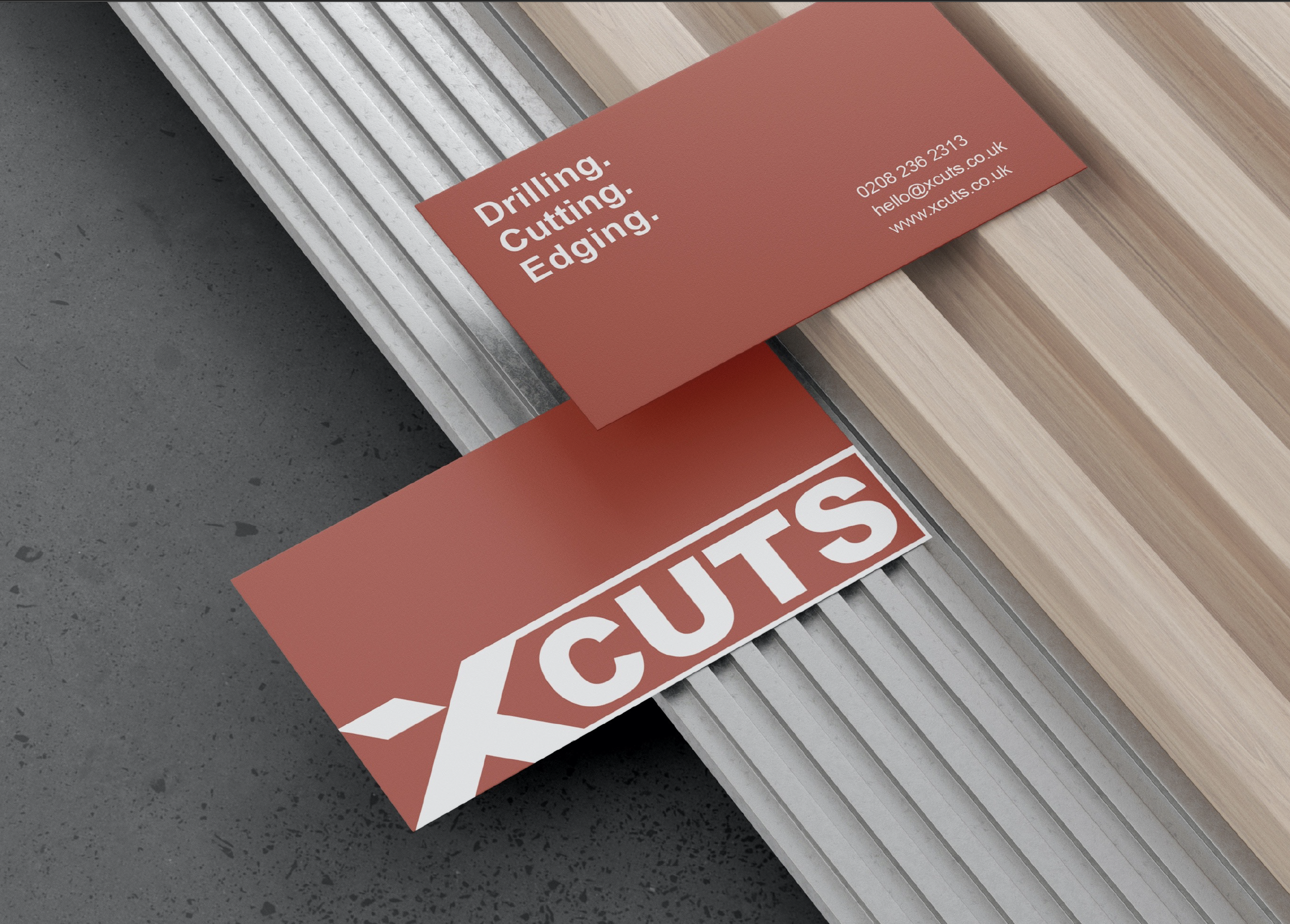 XCuts business cards on a wood and metal slat backdrop — a red Drilling. Cutting. Edging. card with phone, email and web address, and a bold white-on-red XCUTS card